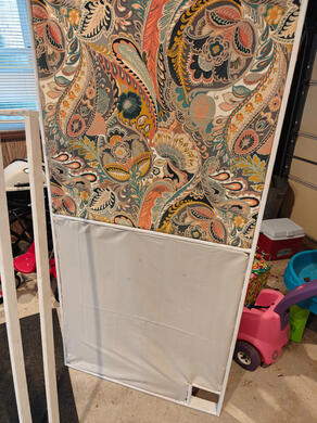 frame with cloth affixed a wooden frame, standing, with paisley cloth stretched across the top half and beige cloth stretched across the bottom half.