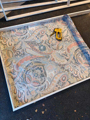 a wooden frame, with paisley cloth laid out, and a staple gun resting on top.
