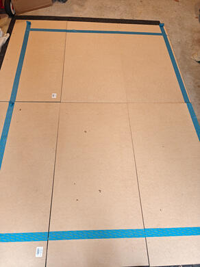 six MDF panels, laid out on the floor. Blue masking tape has been used to measure the dimensions of the wooden flats, for cutting.